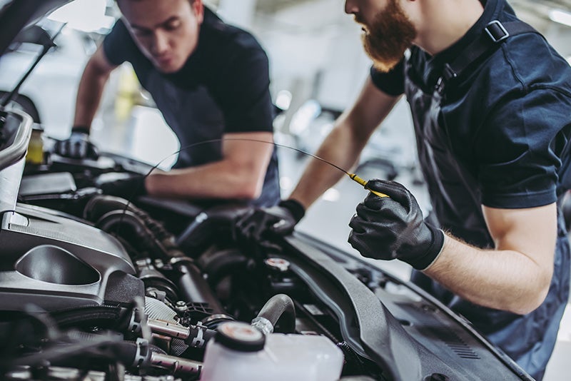 Two auto mechanics working under the hood of a car, with one holding a dipstick to check the engine oil level in a service garage.
